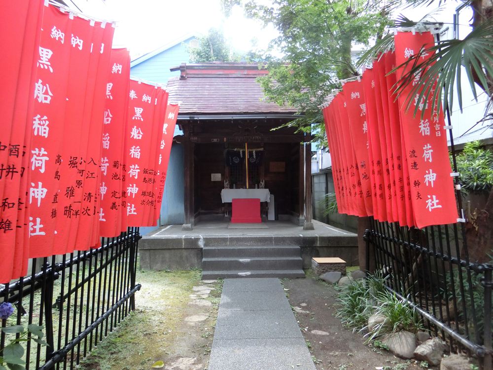 Small shrine with two rows of red flags leading up to it with a blue house and trees in the background.