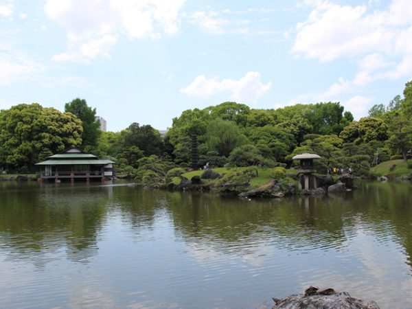 Metropolitan Gardens : 東京都故園, Japanese Culture, The lake at Kiyosumi garden, large tea house, islet with a large Japanese stone lantern, many green trees and a partly cloudy sky in the background.