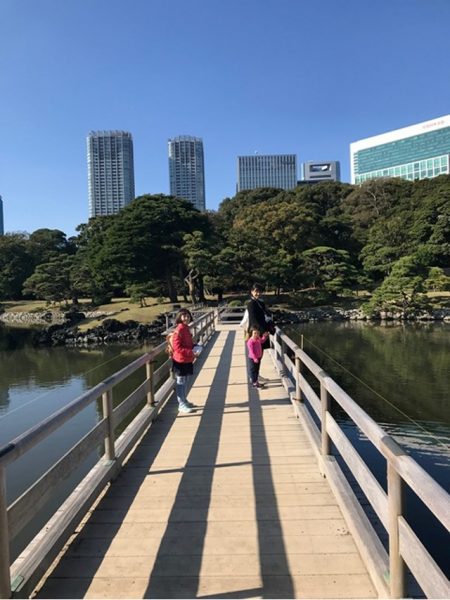 Metropolitan Gardens : 東京都故園, Japanese Culture, A woman and two girls on the wooden walking bridge, green trees, tidal lake and office towers in the background.