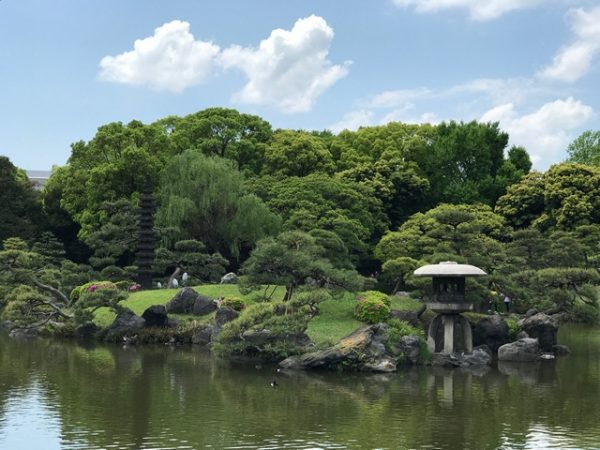 Metropolitan Gardens : 東京都故園, Japanese Culture, Concrete traditional style Japanese lantern on an islet in Kiyosumi gardens lake surrounded by lush trees and grass with a beautiful spring sky sky.