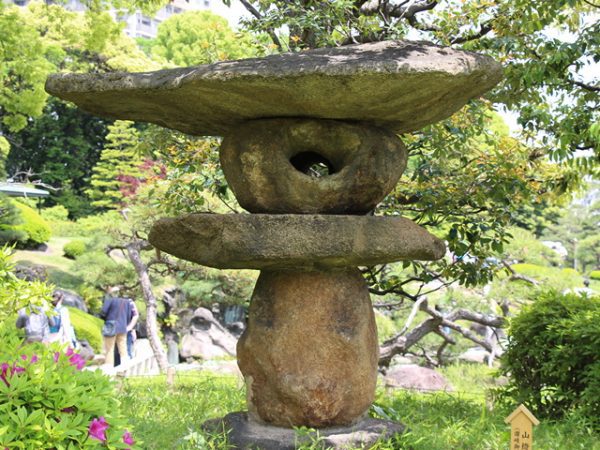 Metropolitan Gardens : 東京都故園, Japanese Culture, Large traditional Japanese stone lantern made of stacked rocks of varying shapes surrounded by trees and shrubbery on a beautiful spring day at Kiyosumi gardens.