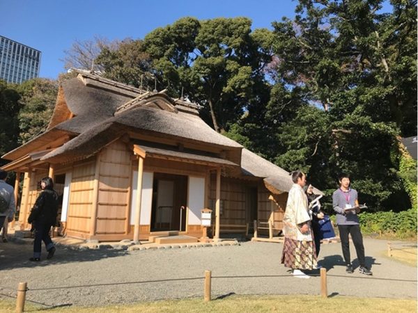 Metropolitan Gardens : 東京都故園, Japanese Culture, A traditional Japanese falcon house, a man in Edo era dress helping some park visitors, green trees, and an office building in the background at Hama-rikyu gardens near downtown Tokyo.