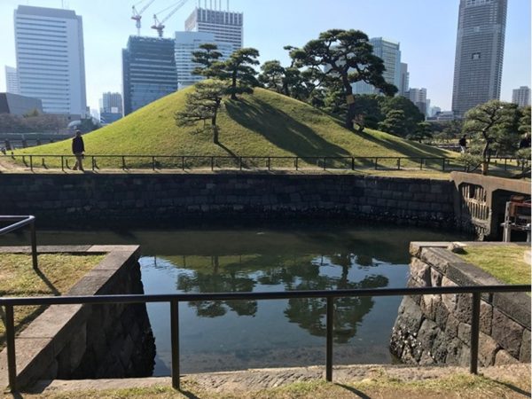 Metropolitan Gardens : 東京都故園, Japanese Culture, An old port area made of stone, a sculpted hill, pine trees, and office towers in the background.