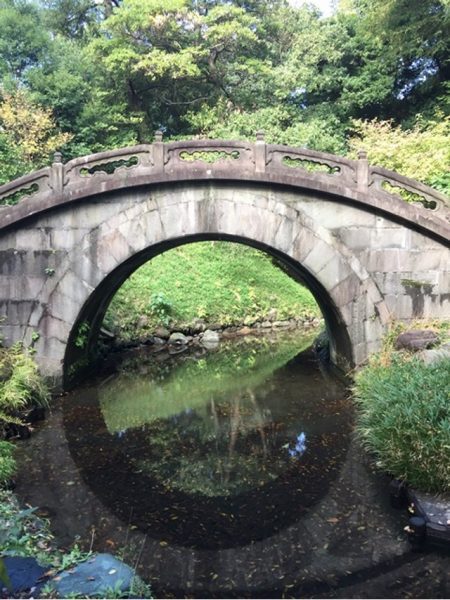 Metropolitan Gardens : 東京都故園, Japanese Culture, Stone Semi-Circle Stone Chinese bridge with a stream running through it, with green and autumn foliage trees in the background.