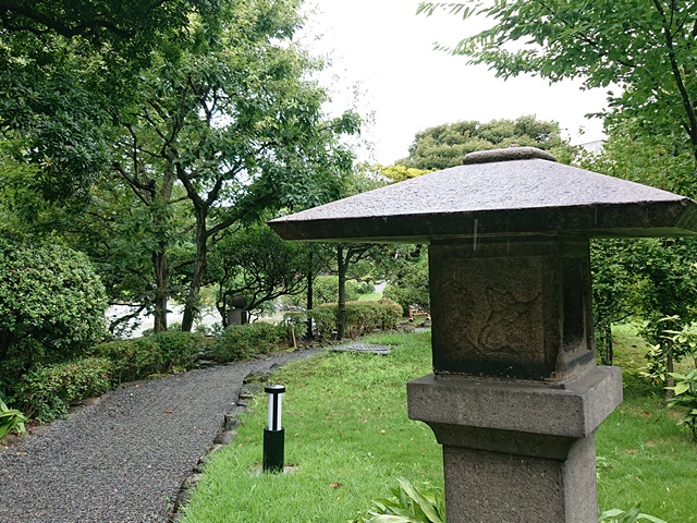 Traditional Japanese stone lantern and walkway through the garden with green trees, shrubs, and grass.