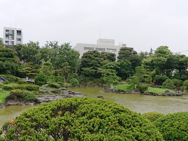 The tidal pond, some islets, large unusual stones along the bank, many trees and shrubs and some office buildings in the background at Kyu-Yasuda Garden in Sumida City of Tokyo.