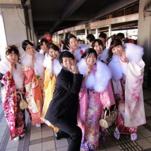 Several young women wearing colorful kimonos and a young man in a black suit are posing for a photo in front of a city hall in Japan.