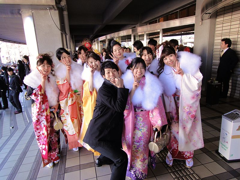 Several young women wearing colorful kimonos and a young man in a black suit are posing for a photo in front of a city hall in Japan.