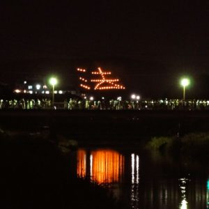 A mountainside in Kyoto set on fire in the shape of a kanji character that is also reflected on the river with onlookers all around.