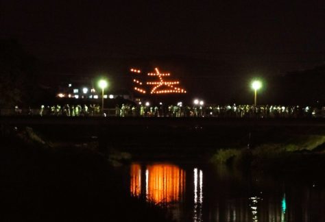A mountainside in Kyoto set on fire in the shape of a kanji character that is also reflected on the river with onlookers all around.