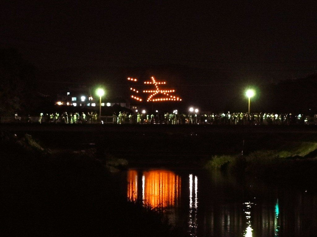 A mountainside in Kyoto set on fire in the shape of a kanji character that is also reflected on the river with onlookers all around.