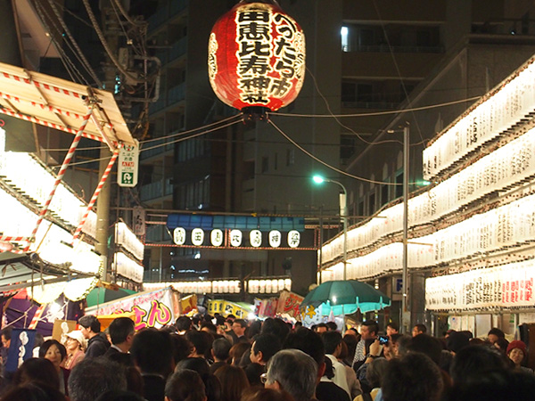 A huge red Japanese traditional lantern hanging high up flanked by rows of white lanterns with a huge crowd of festival goers beneath.